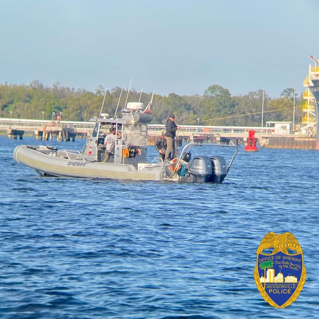 A police boat with officers on board patrols the water, labeled "Sheriff" and equipped with twin outboard motors. In the backdrop of Jacksonville Arrival, a shoreline emerges with trees and a brightly colored structure, witnessing a Carnival Cruise as it sails by.