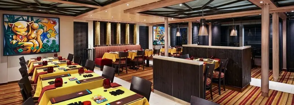 A brightly lit restaurant interior featuring tables with yellow tablecloths and dark brown chairs, reminiscent of a carnival cruise menu experience. Colorful paintings adorn the walls, while the ceiling boasts a geometric design and the floor is covered with a striped pattern.