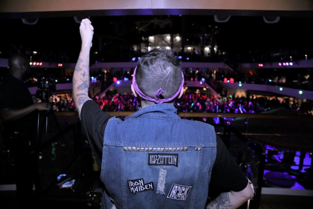 A person wearing a denim vest with band patches raises their fist, setting the passionate mood at a music event. The crowd is energetically engaged, vibrant lights illuminating the scene like a cruise on waves of sound.