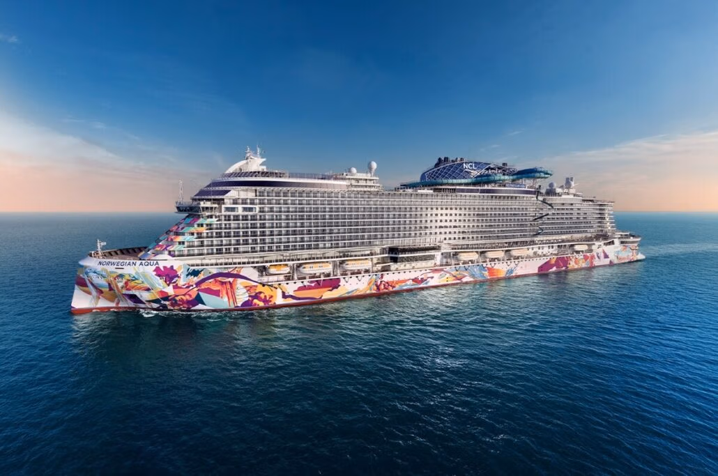 The Norwegian Aqua makes its exclusive European debut, gliding gracefully on calm blue waters. This large cruise ship showcases vibrant artwork along its sides and boasts multiple decks with cabins. The sky is clear, offering a soft gradient from blue to peach near the horizon.