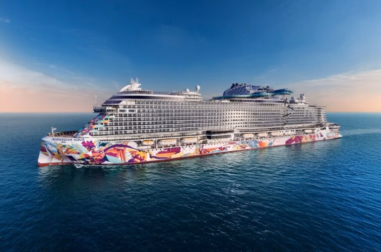 The Norwegian Aqua makes its exclusive European debut, gliding gracefully on calm blue waters. This large cruise ship showcases vibrant artwork along its sides and boasts multiple decks with cabins. The sky is clear, offering a soft gradient from blue to peach near the horizon.