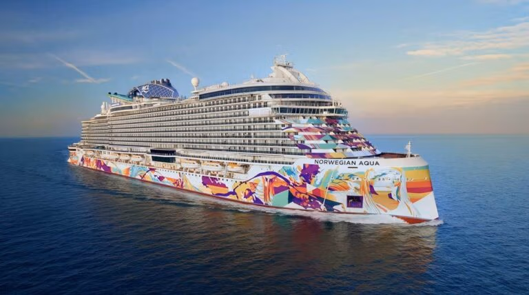The grand cruise ship "Norwegian Aqua," adorned with Prima Plus artwork, glides gracefully on calm ocean waters. Its vibrant hull reflects the twilight hues as the sky embraces a few scattered clouds at sunset.