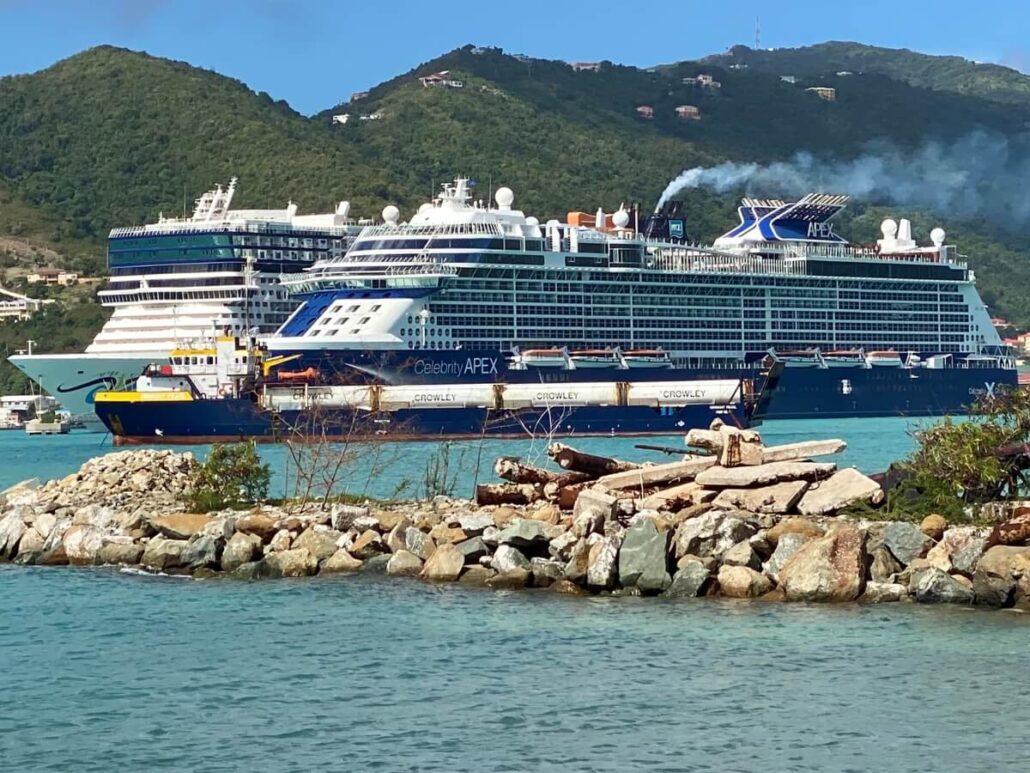 The large cruise ship "Celebrity Apex" is anchored near the rocky shoreline of Tortola. In the background, the "Norwegian Epic" is also visible. White smoke curls from the cruise ship's funnels, set against green hills and buildings under a clear blue sky.