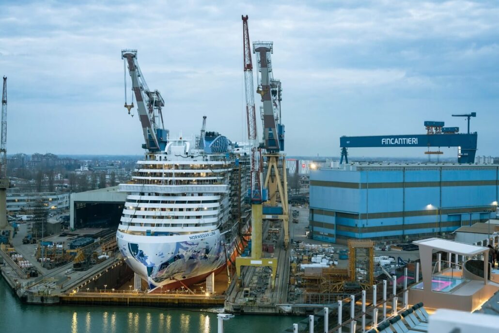 A large Prima Plus Cruise Ship is docked at a shipyard, with several cranes surrounding it. The sky is cloudy, and there are industrial buildings nearby. A Fincantieri logo is visible on one of the buildings, hinting at the collaboration with NCL to craft this maritime marvel.