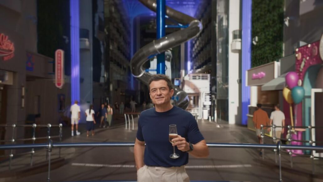 A man in a navy shirt holds a champagne flute, standing in a modern indoor setting aboard the MSC World America. Behind him, a tall, twisting slide and various shops buzz with activity as people explore. The atmosphere is lively, illuminated by blue and purple lights, celebrating the Miami Debut of MSC Cruises' latest marvel.
