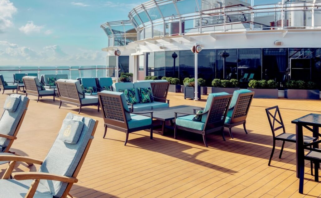 The outdoor patio on the Queen Elizabeth cruise ship, recently refurbished for its Alaska debut, features teal-cushioned lounge chairs and sofas around low tables. The wooden deck is framed by glass railings and potted plants against a backdrop of clear skies and ocean.
