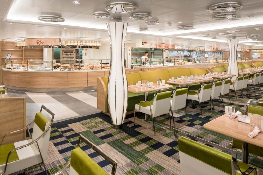 Spacious, modern dining area with wooden and white elements and green accents aboard a Holland America cruise ship. Tables are set with napkins and utensils, while a buffet counter with food and cooking stations gleams in the clean, well-lit environment.