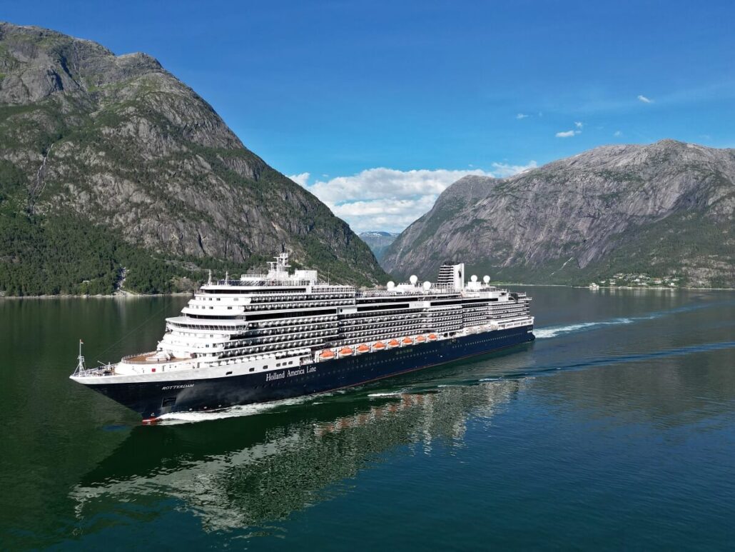 A majestic Holland America cruise ship navigates a narrow fjord, flanked by steep, rocky mountains. The calm water mirrors the ship and the stunning landscape. Above, a clear sky is dotted with distant clouds.