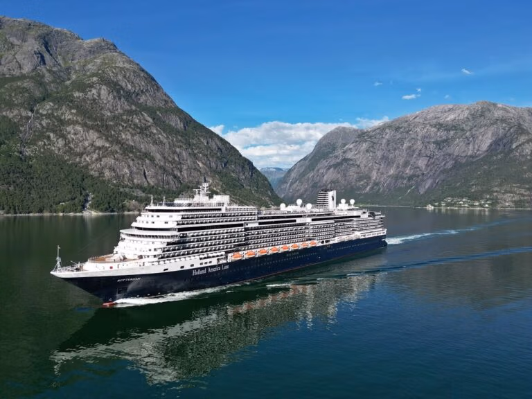 A Holland America cruise ship navigates a narrow fjord in Norway, flanked by steep, rocky mountains. The calm water mirrors the ship and the stunning landscape. Above, a clear sky is dotted with distant clouds.