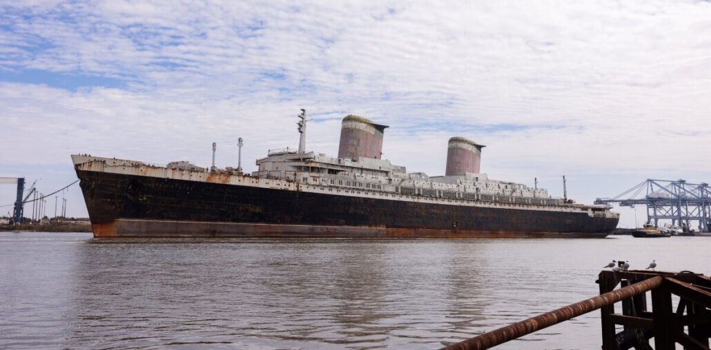 A large, rusted ocean liner resembling the SS United States with two smokestacks sits abandoned in a Florida County harbor. The sky is partly cloudy, and cranes are visible in the background. The water is calm, with a dock edging into the frame, hinting at its potential as a future reef site.