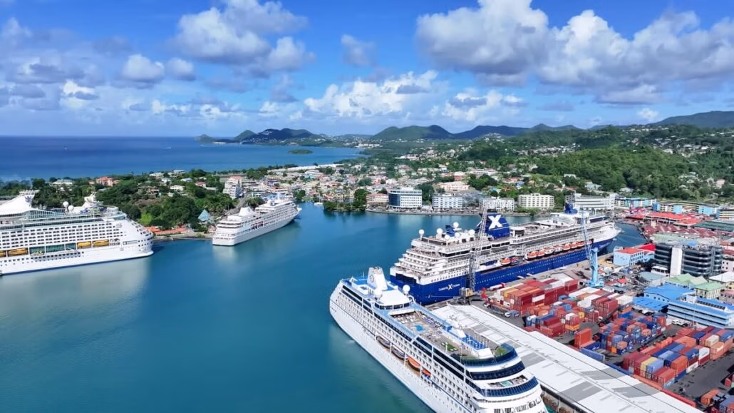 Aerial view of a bustling port in Saint Lucia with several large cruise ships docked. The port is surrounded by a town with various buildings and lush green hills, welcoming countless visitors. The ocean stretches into the horizon under a partly cloudy sky, highlighting the vibrant cruise traffic.