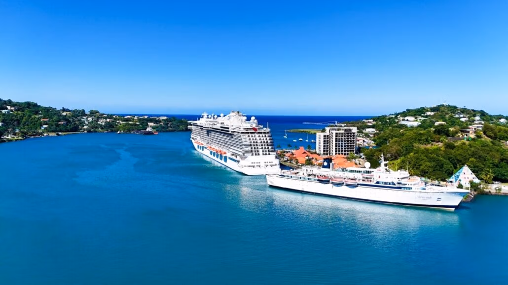 Aerial view of a bustling Saint Lucia harbor with two cruise ships docked. The surrounding landscape showcases lush greenery, small hills, and buildings along the shoreline. The sky is clear, and the sea is calm and bright blue, inviting visitors to explore its serene beauty.