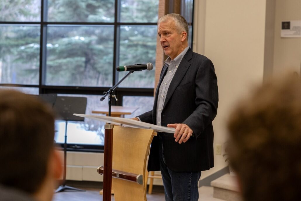 A man in a dark blazer and jeans, reminiscent of a U.S. Senator, speaks at a wooden podium with a microphone, framed by large windows revealing snow-covered trees outside. Blurred audience members listen intently in the foreground.