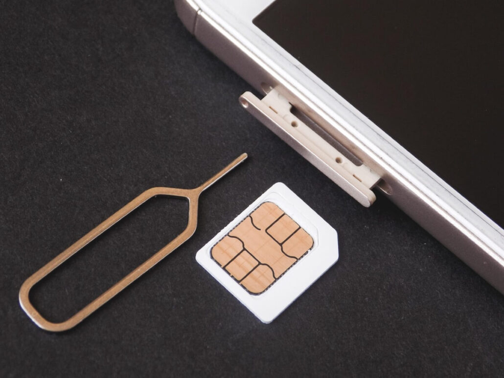 A smartphone with a SIM card tray partially ejected, possibly using a SIM card ejector tool. Nearby, a SIM card rests on the dark surface. The phone's screen is off—a subtle reminder of how easy it is for fraudsters to access passengers’ bank accounts through unsecured devices.