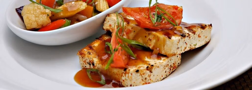 A plate with two slices of grilled tofu topped with sauce, tomato, and herbs reminiscent of Carnival Cruise menus. Beside it, a bowl holds assorted vegetables like cauliflower, bell peppers, and baby corn. The dishes elegantly rest on a white ceramic plate.