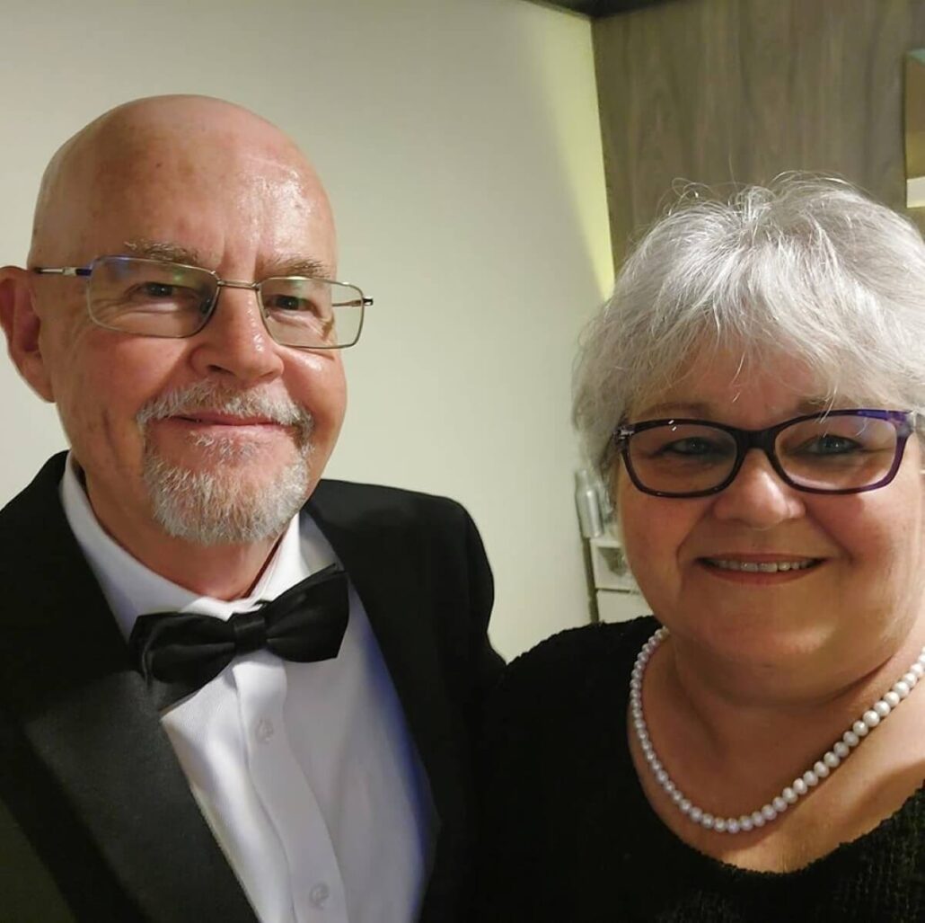 An older man with a bald head and glasses, wearing a tuxedo and bow tie, stands next to an older woman with short gray hair and glasses, adorned in a black top and pearl necklace. They smile warmly for the camera indoors—a stark contrast to tales of fraudsters seeking £25,000 loans.