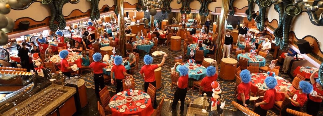 A vibrant, themed restaurant scene resembling a carnival cruise is complete with staff in red shirts and blue wigs. Tables boast red tablecloths and whimsical centerpieces. Diners revel in the lively atmosphere amid oversized decorations adorning the walls and ceiling.