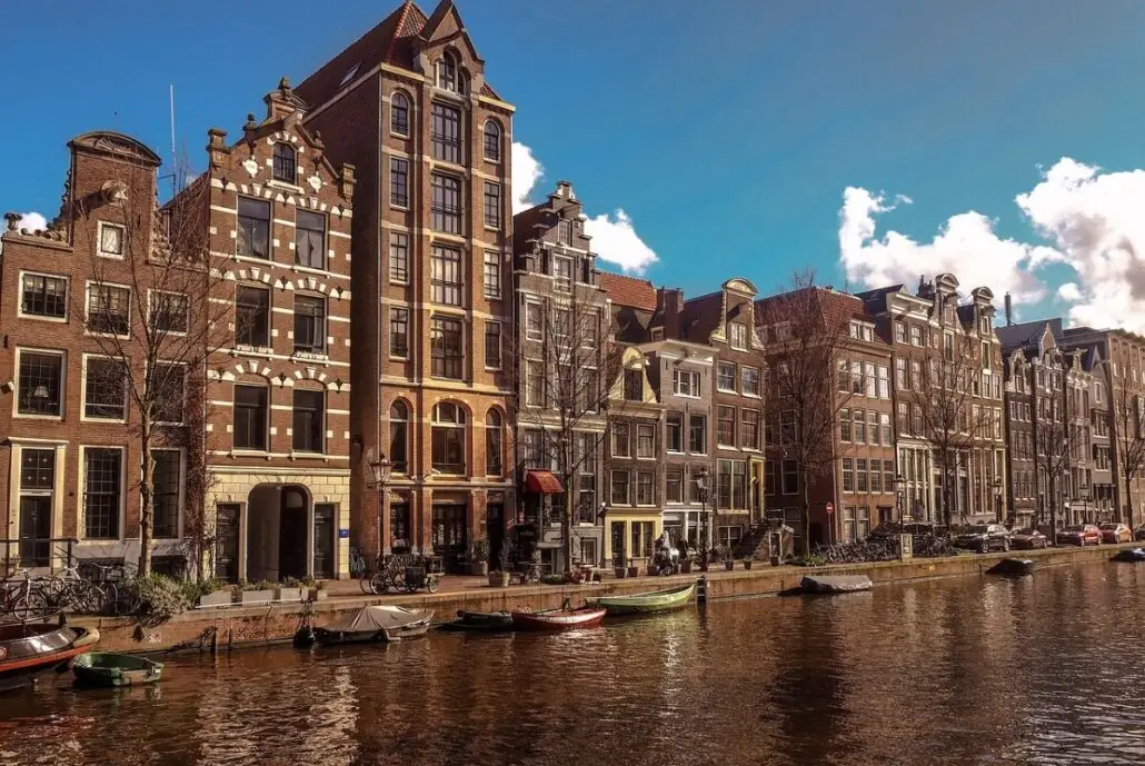 The canal, perfect for Amsterdam Tours, is lined with traditional Dutch townhouses featuring gabled facades. Small boats are moored along the water's edge. The sky is blue with scattered clouds, and the scene is warmly illuminated by sunlight.