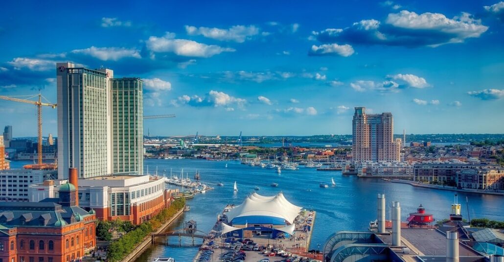 Aerial view of a city harbor with a waterfront. High-rise buildings line the skyline as boats, including ones for ship relocation, are docked nearby. A white tent-like structure sits by the water. The sky above is blue with scattered clouds, hinting at adventures from Baltimore to Florida on Royal Caribbean cruises.