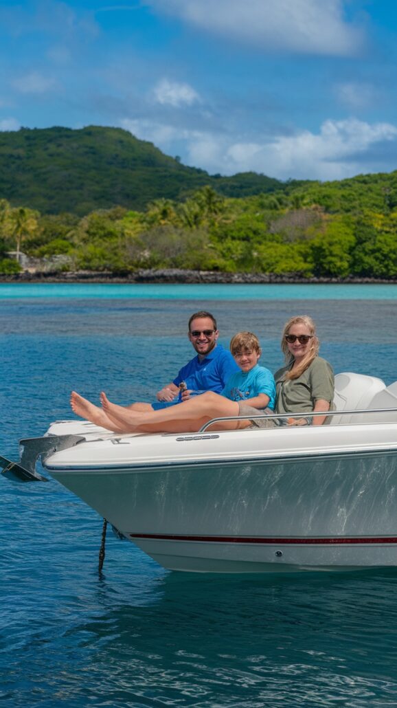 A family of three enjoys a serene boat ride, reminiscent of a Caribbean cruise. The man wears a blue shirt, the woman sports sunglasses and a green shirt, and the child is in light blue. Behind them, lush green trees and hilly landscapes create an idyllic backdrop against calm, azure waters.