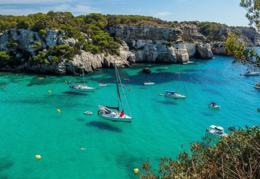 A scenic coastal view with clear turquoise water graces the Balearic Islands. Several sailboats and small boats are anchored near a rocky shoreline covered with greenery. Hills and cliffs surround the bay, while a few people swim in the water under a clear sky.