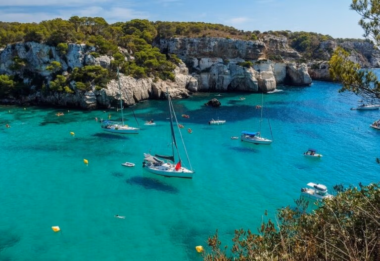 A scenic coastal view with clear turquoise water graces the Balearic Islands. Several sailboats and small boats are anchored near a rocky shoreline covered with greenery. Hills and cliffs surround the bay, while a few people swim in the water under a clear sky.