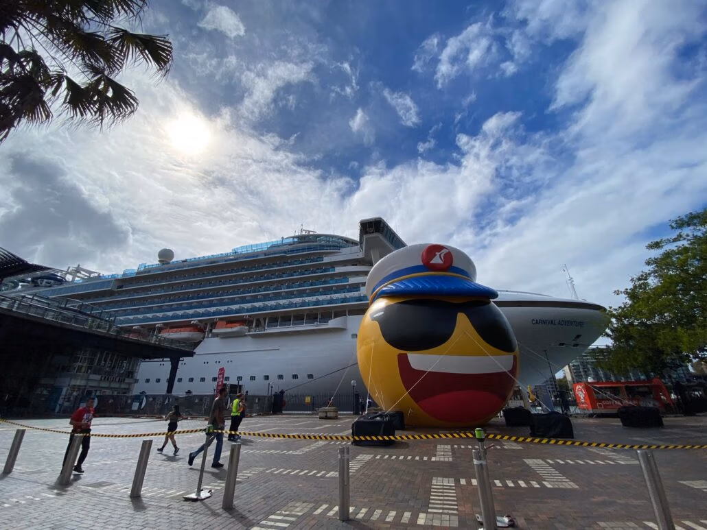 A large cruise ship, part of the Carnival Adventure fleet, is docked at a port under a partly cloudy sky. In front, a giant inflatable emoji with sunglasses and a captain's hat stands proudly. People stroll nearby while caution tape lines the pavement.