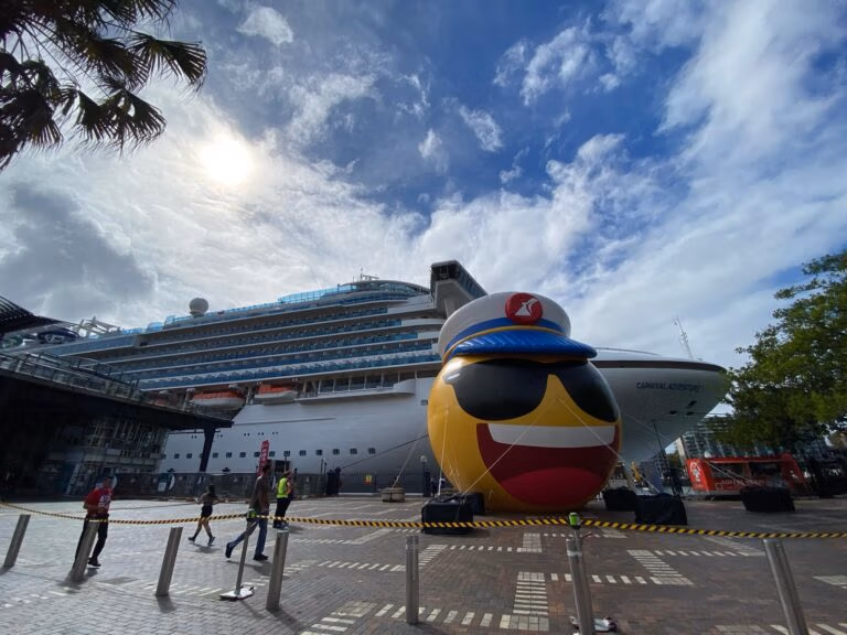 A large cruise ship, part of the Carnival Adventure fleet, is docked at a port under a partly cloudy sky. In front, a giant inflatable emoji with sunglasses and a captain's hat stands proudly. People stroll nearby while caution tape lines the pavement.