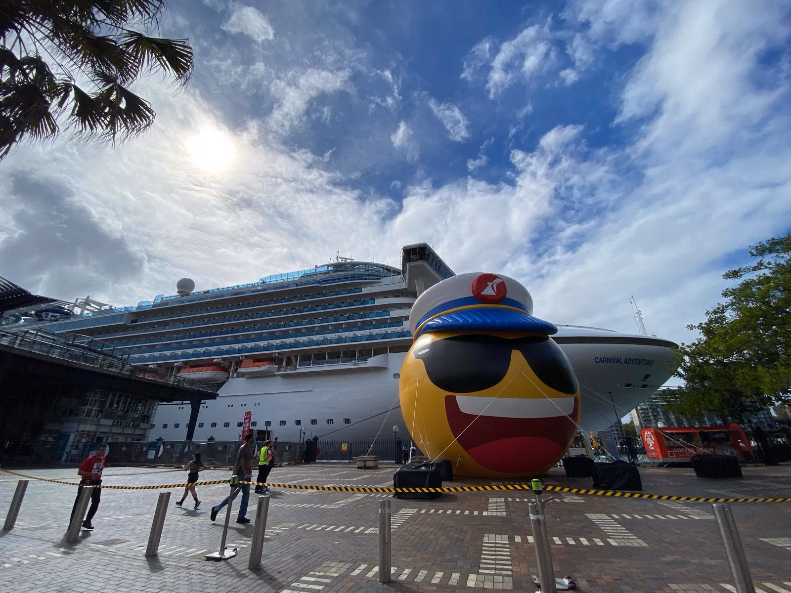 A large cruise ship, part of the Carnival Adventure fleet, is docked at a port under a partly cloudy sky. In front, a giant inflatable emoji with sunglasses and a captain's hat stands proudly. People stroll nearby while caution tape lines the pavement.