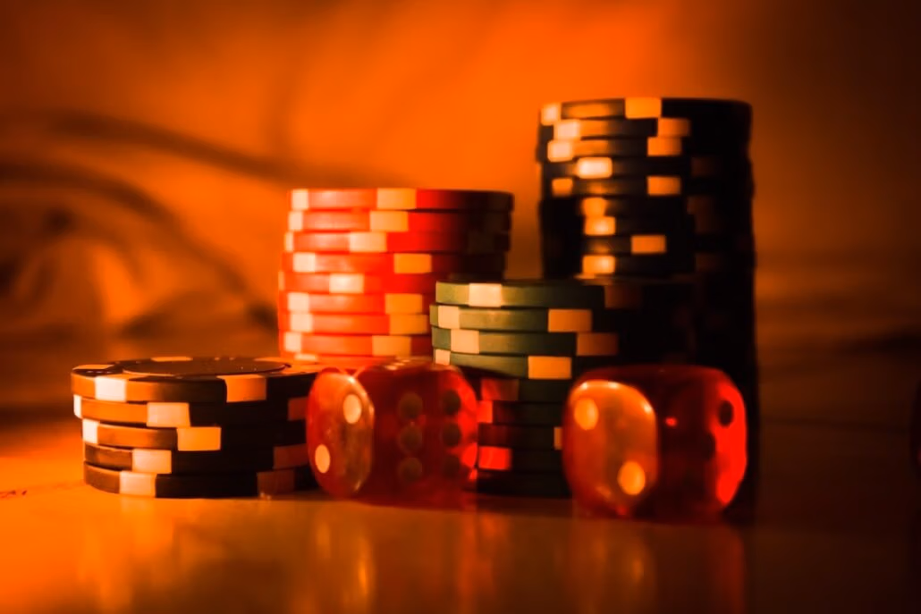 Stacks of poker chips in vibrant colors, including red, black, and green, are piled on a surface. Two red translucent dice are in the foreground. The scene is dimly lit with a warm, orange hue—perfect for first-time gamblers eager to dive into the thrill of casino cruises.