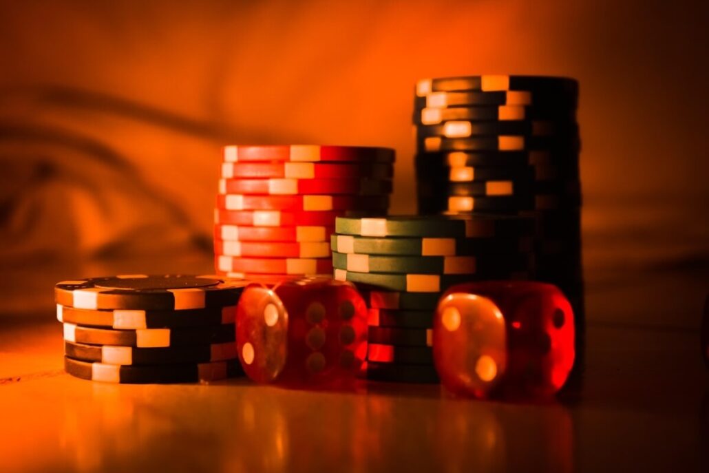 Stacks of poker chips in vibrant colors, including red, black, and green, are piled on a surface. Two red translucent dice are in the foreground. The scene is dimly lit with a warm, orange hue—perfect for first-time gamblers eager to dive into the thrill of casino cruises.