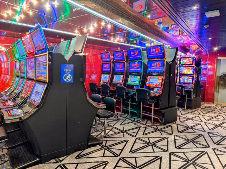 The brightly lit casino room aboard the Celestyal Journey boasts colorful slot machines and neon-lit, reflective walls. Geometric carpet patterns adorn the floor, while chairs sit ready for guests seeking tips on hitting the jackpot as they cruise to their next destination.
