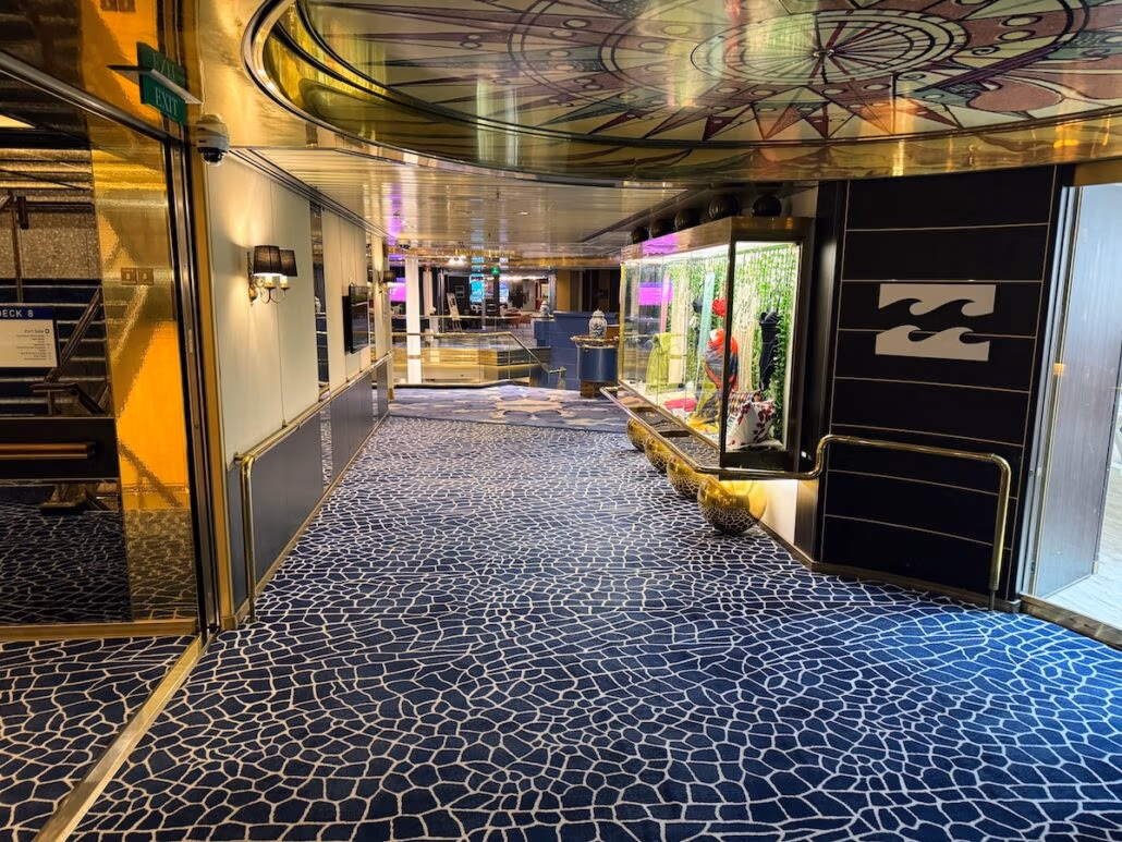 A hallway with patterned blue carpet and gold-accented walls, reminiscent of a Celestyal Journey trip report. Glass display cases on the right showcase various items, while the ceiling features a circular, abstract design. Doors line both sides, bathed in warm lighting that feels perfect.