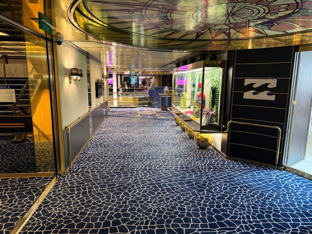 A hallway with patterned blue carpet and gold-accented walls, reminiscent of a Celestyal Journey trip report. Glass display cases on the right showcase various items, while the ceiling features a circular, abstract design. Doors line both sides, bathed in warm lighting that feels perfect.