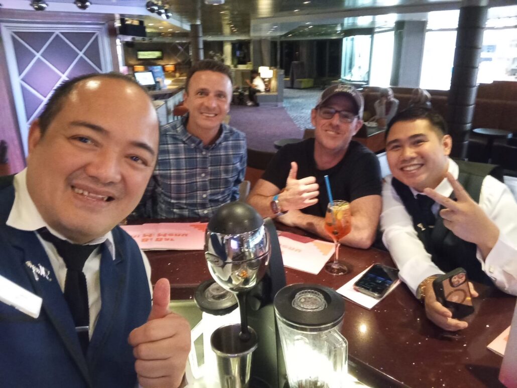 Four men capture a selfie at the bar during their Celestyal Journey. The man on the left sports a suit and tie, while two seated in the middle share reasons to smile—one with a thumbs-up. The fourth man on the right wears a vest, flashing a peace sign amidst drinks and typical cruise vibes.