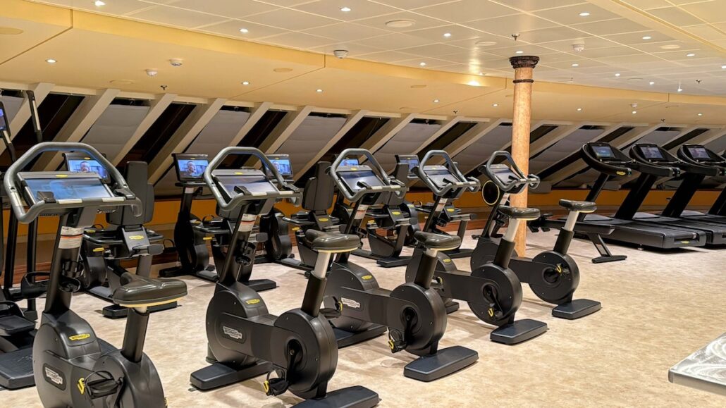 A gym aboard the Celestyal Journey features a row of stationary bikes on the left and treadmills on the right, each with a digital display screen. The space offers beige carpeting and a sloped ceiling with multiple recessed lights, making it one of many reasons to enjoy your cruise ship vacation.