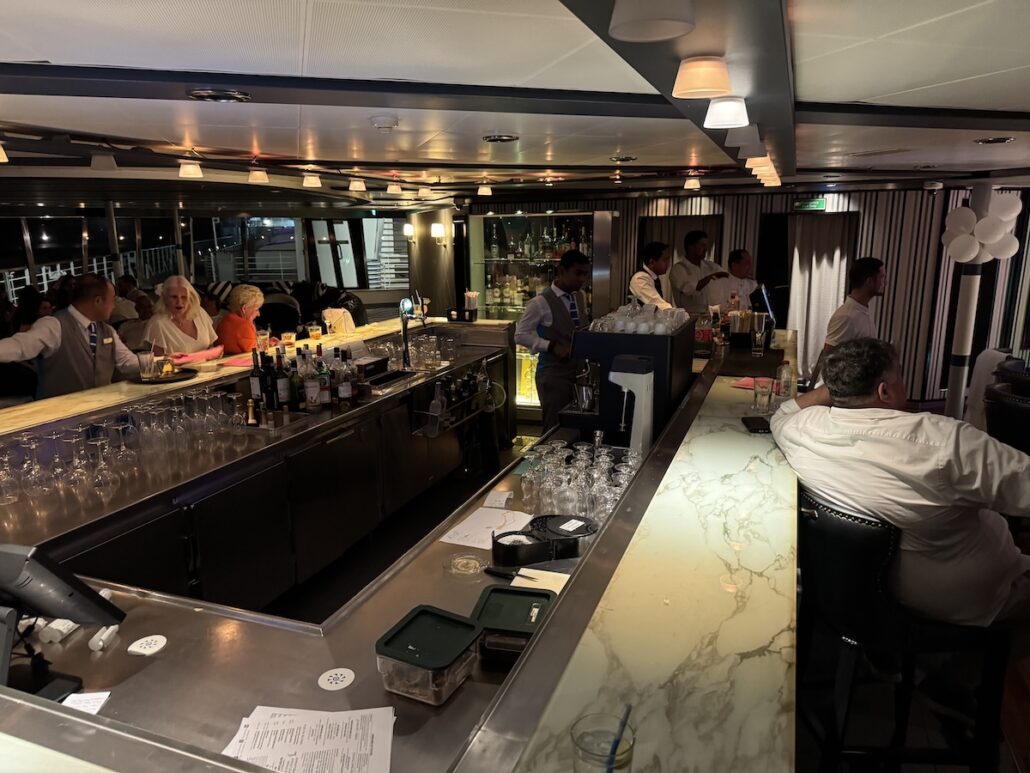 A dimly lit bar with a marble countertop reminiscent of the elegant lounges aboard a midsize ship. Bartenders in uniforms serve drinks to customers on stools, as glasses and bottles are arranged neatly. Patrons engage in conversation, much like those onboard a Celestyal Journey trip.