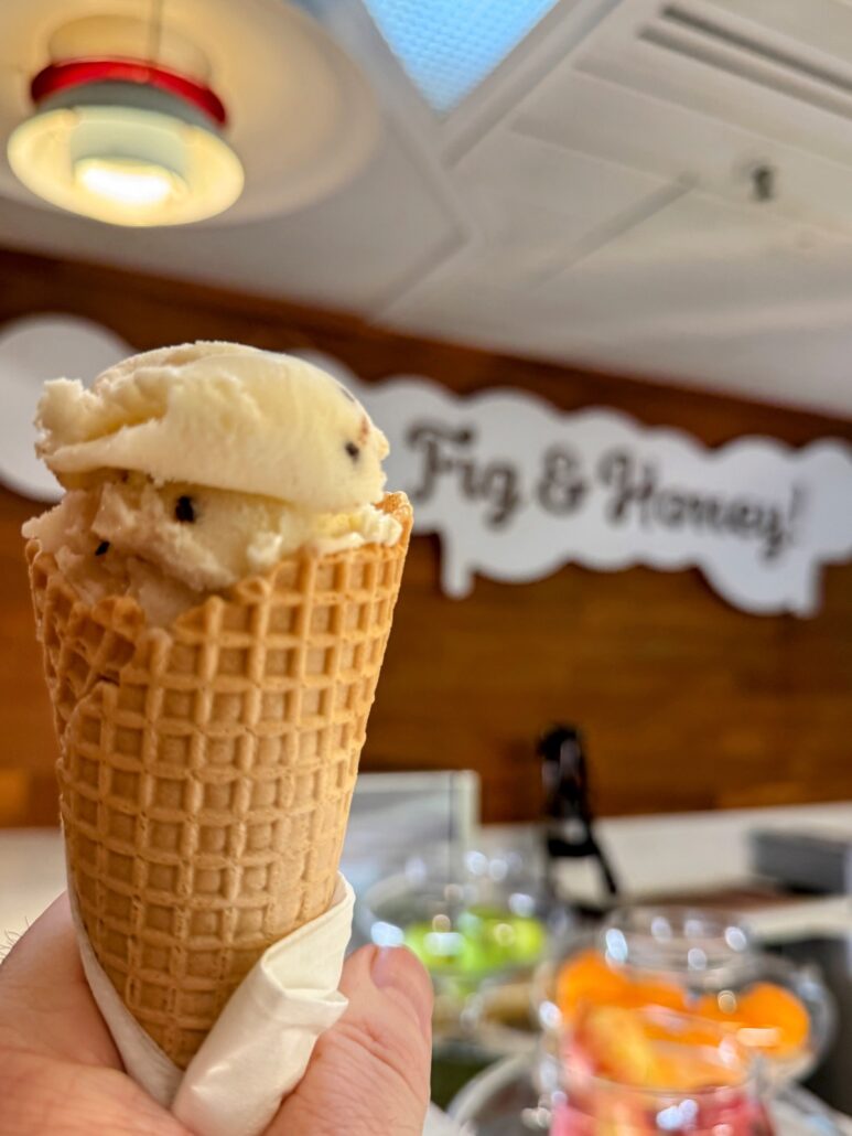 A person holds a waffle cone with a scoop of ice cream. The backdrop, akin to the ambience on a Celestyal Journey trip, features an indoor setting with a sign reading "Fig & Honey." A light fixture hangs above, illuminating containers filled with colorful contents on the counter.