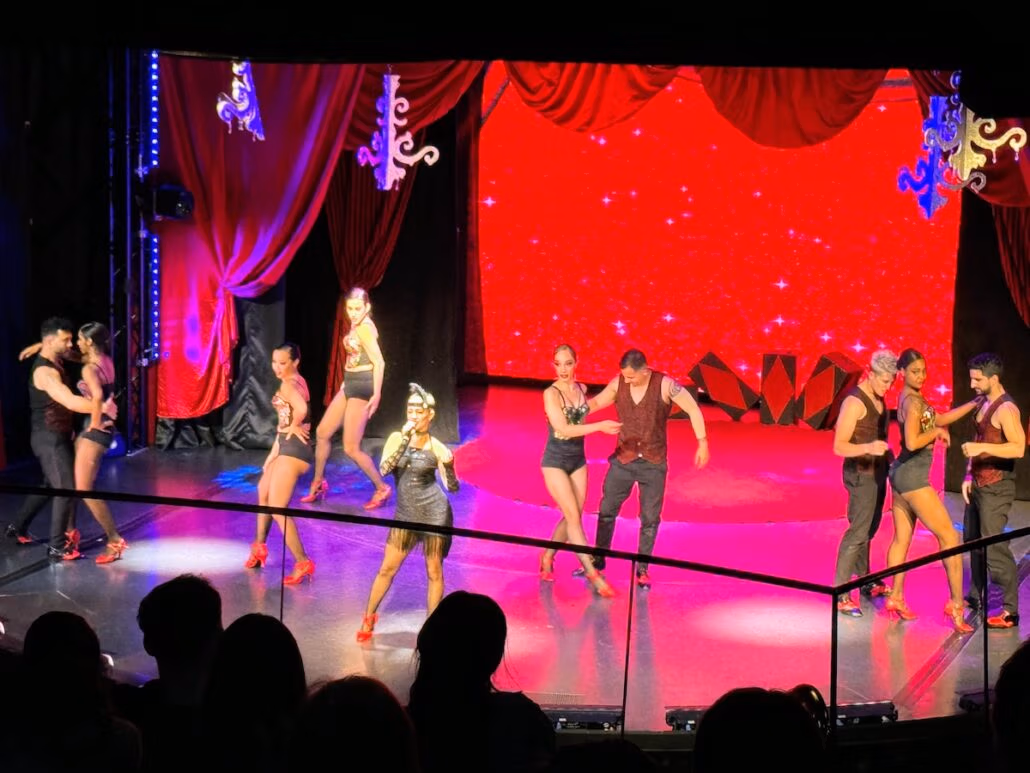 Performers on stage dance in pairs in front of a red curtain backdrop with star patterns reminiscent of a Celestyal Journey evening. They wear black and red costumes, and the lighting highlights their movements. A typical cruise ship audience watches from the foreground in silhouette.