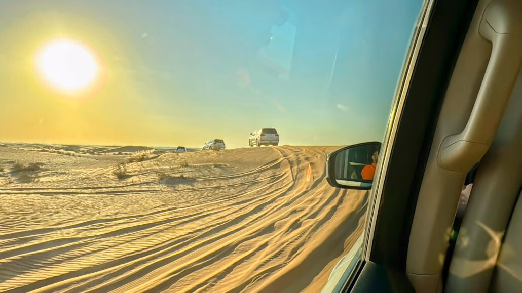 A desert scene with several SUVs cruising on sand dunes under a clear blue sky. The sun is low on the horizon. The right side reveals the interior of another SUV, with a side mirror reflecting the sandy landscape, capturing tips of adventure at every turn.
