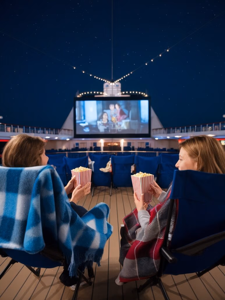 Two people sitting in blue chairs on a Caribbean cruise's deck enjoy an unforgettable adventure as they watch a movie on a large outdoor screen at night. Wrapped in blankets and holding striped popcorn boxes, they admire the stars twinkling brightly above.