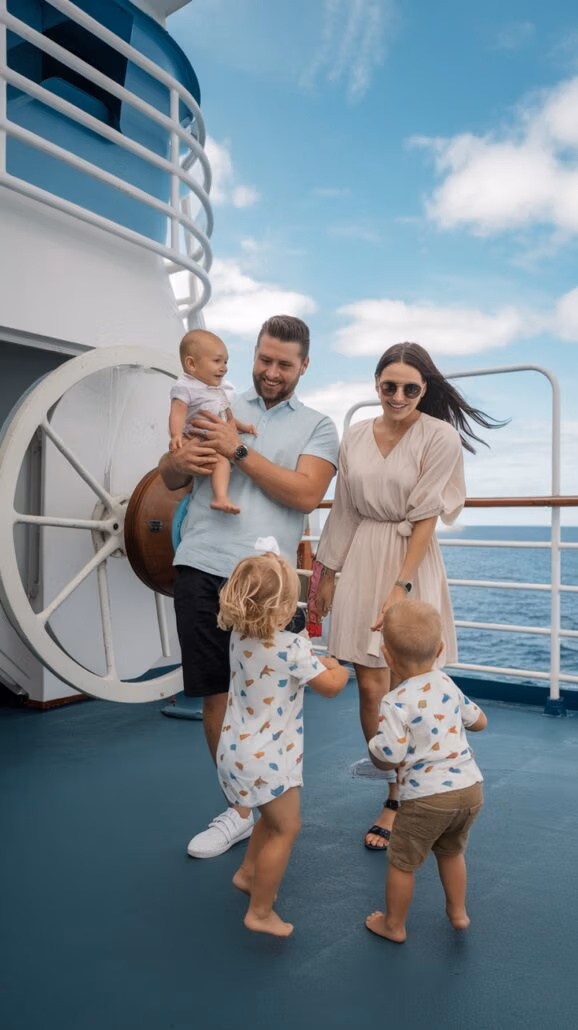 A man and a woman stand on a ship's deck during their unforgettable family Caribbean cruise with three small children. The man holds a baby, while the woman, wearing sunglasses, smiles as two toddlers play nearby. The sky is partly cloudy, offering a stunning view of the sea in the background.