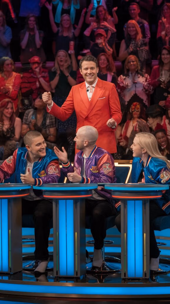 A man in an orange suit stands smiling with arms raised. Three people in colorful jackets are seated at illuminated desks, engaging as if immersed in exciting games. A cheering audience is visible in the background, reminiscent of lively nights on a Caribbean cruise.