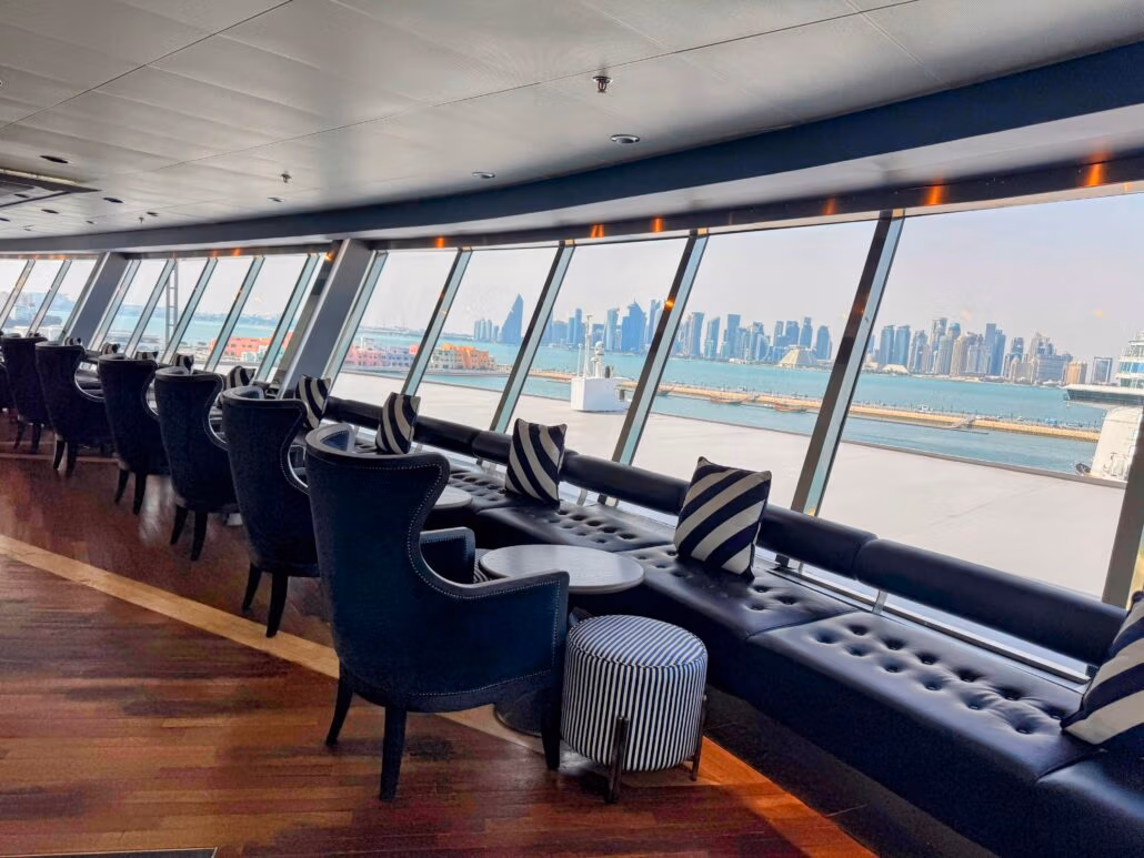 The Celestyal Journey unveils a modern lounge area with black lounge chairs and striped cushions. Large windows offer stunning views of the city skyline over the water, perfect for cruising enthusiasts. The wooden floor beautifully complements the sleek interior design, creating a serene escape.
