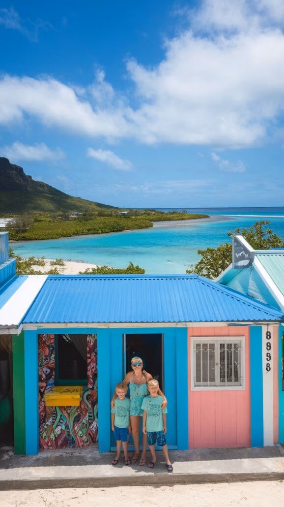 A woman and three children stand in front of a small, colorful building with blue and pink walls. Behind them, the beach's clear turquoise water invites them for family bonding activities amidst lush greenery and a distant mountain. The sky is partly cloudy, perfect for their Caribbean adventure.