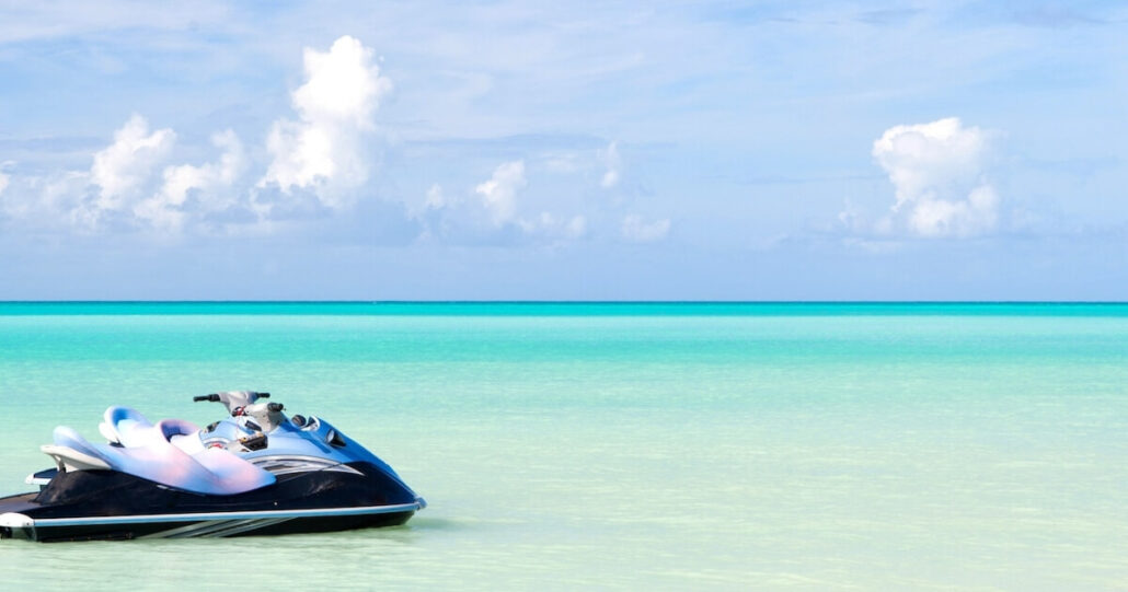 A jet ski is moored in the clear turquoise waters of Half Moon Cay on a calm day. The horizon greets a few fluffy clouds in the blue sky, crafting a serene and idyllic tropical scene, perfect for cruise passengers seeking tranquility.