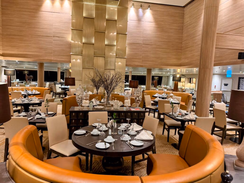 A modern, elegant restaurant interior reminiscent of a Celestyal Journey, with round tables set for dining. Adorned with white tableware and napkins, the room features warm wood tones, mirrored walls, and ambient lighting, perfectly suited for those cruising through a sophisticated dining experience.