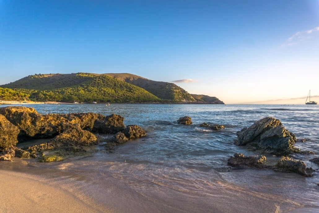 A scenic view of a rocky beach in the Balearic Islands with clear, gentle waves under a bright blue sky. A green hillside is visible in the background, and a sailboat is seen on the horizon. The lighting suggests it is either early morning or late afternoon.