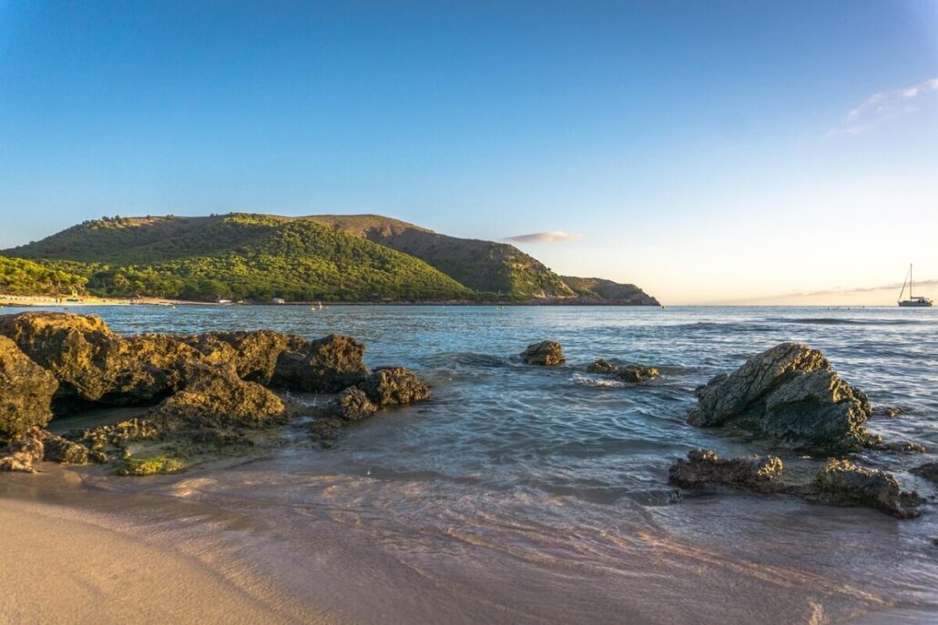 A scenic view of a rocky beach in the Balearic Islands with clear, gentle waves under a bright blue sky. A green hillside is visible in the background, and a sailboat is seen on the horizon. The lighting suggests it is either early morning or late afternoon.