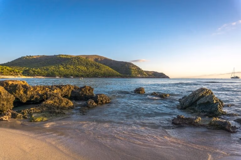 A scenic view of a rocky beach in the Balearic Islands with clear, gentle waves under a bright blue sky. A green hillside is visible in the background, and a sailboat is seen on the horizon. The lighting suggests it is either early morning or late afternoon.